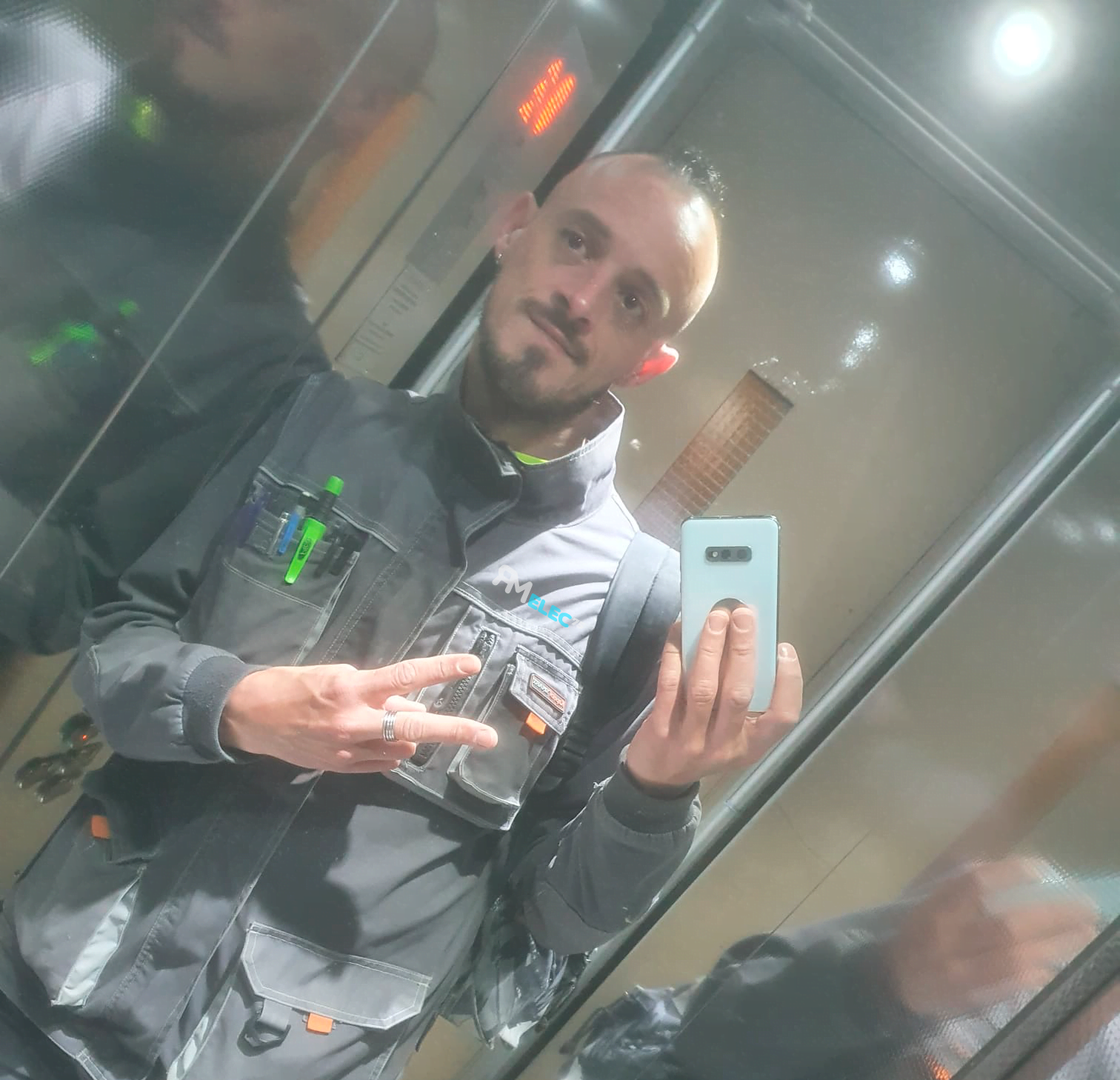 Technicien en uniforme gris dans un ascenseur tenant un smartphone et faisant un signe de paix.